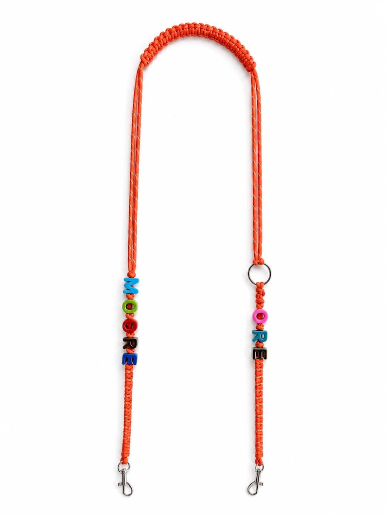 Op de afbeelding: Een oranje gevlochten koord met twee zilverkleurige metalen karabijnsluitingen aan de uiteinden. E&eacute;n kant bevat kleurrijke letterkralen die MOORE spellen, terwijl de andere kant letterkralen met ORE en een zilverkleurige metalen ring bevat.