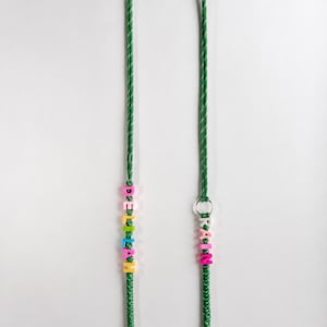Op de afbeelding: Een groen gevlochten koord met twee secties van kleurrijke plastic letterkralen. De linkerkant spelt DELILAH en de rechterkant spelt RAIN. Het koord bevat een zilverkleurige metalen ring en twee zilverkleurige metalen draaibare karabijnsluitingen.
