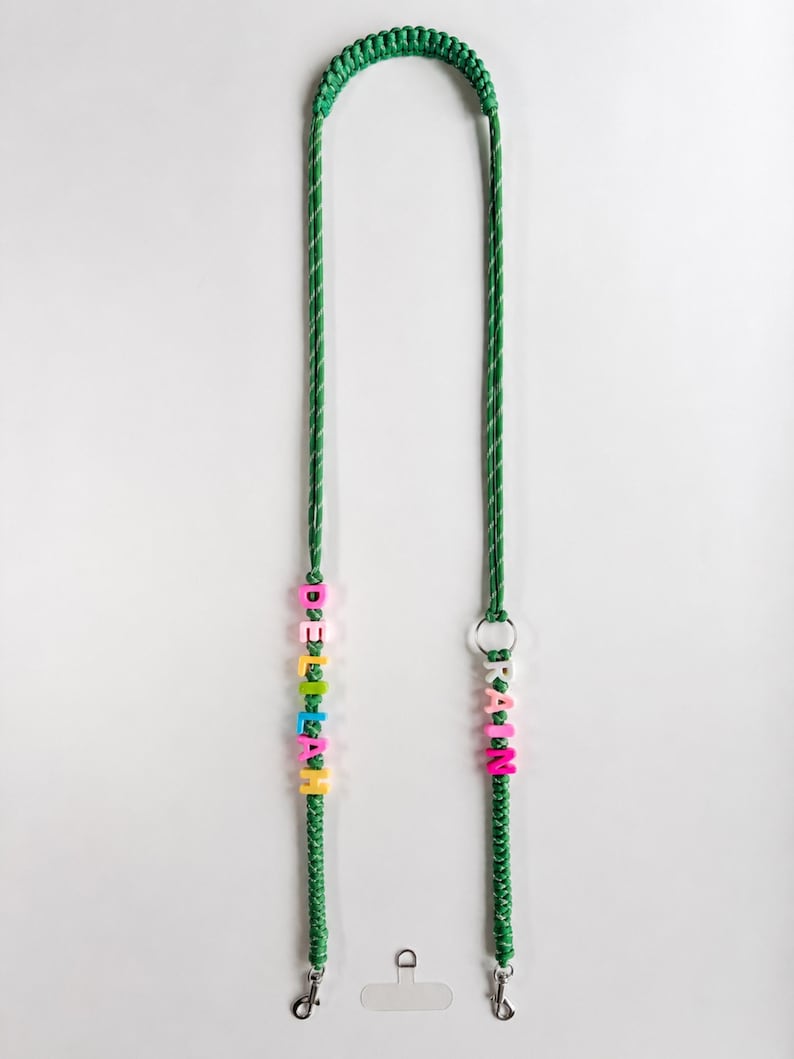 Op de afbeelding: Een groen gevlochten koord met twee secties van kleurrijke plastic letterkralen. De linkerkant spelt DELILAH en de rechterkant spelt RAIN. Het koord bevat een zilverkleurige metalen ring en twee zilverkleurige metalen draaibare karabijnsluitingen.