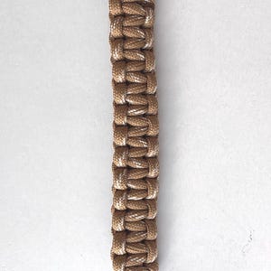 May include: Tan and white braided paracord with a curved top section and two parallel strands. The paracord is a neutral color, suitable for various crafting projects or accessories. The image is set against a plain white background.