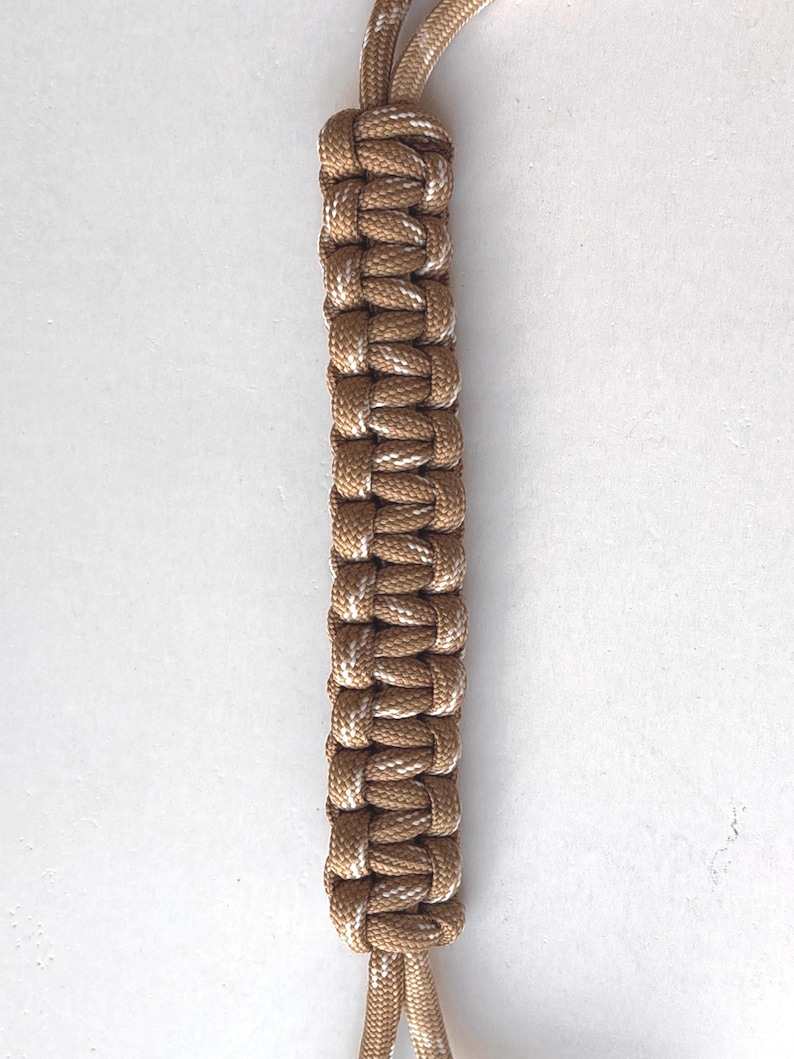 May include: Tan and white braided paracord with a curved top section and two parallel strands. The paracord is a neutral color, suitable for various crafting projects or accessories. The image is set against a plain white background.
