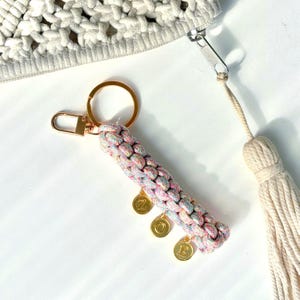 Puede incluir: Un llavero con un cordón trenzado en un patrón multicolor pastel, unido a una anilla y un cierre de metal dorado. Tres pequeños dijes circulares dorados cuelgan del cordón, cada uno grabado con las letras Z, O y E.