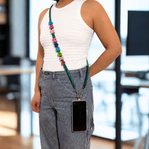 Op de afbeelding: Een crossbody telefoonkoord bestaande uit een donkergroen gevlochten koord versierd met kleurrijke letterkralen die het woord "SUMMORO" vormen. Het koord wordt met metalen clips aan een telefoonhoesje bevestigd om het toestel aan de heup te dragen.