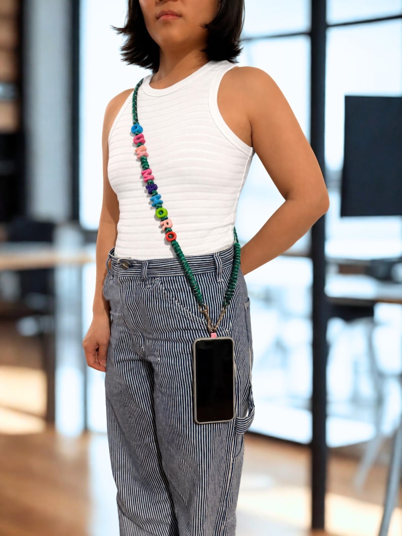 Op de afbeelding: Een crossbody telefoonkoord bestaande uit een donkergroen gevlochten koord versierd met kleurrijke letterkralen die het woord "SUMMORO" vormen. Het koord wordt met metalen clips aan een telefoonhoesje bevestigd om het toestel aan de heup te dragen.