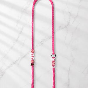Op de afbeelding: Een felroze gevlochten koord met witte en roze letterkralen die SOCIA en EMI spellen. Het koord is voorzien van een zwarte ring en twee zilverkleurige metalen karabijnsluitingen aan de onderkant voor een veilige bevestiging van voorwerpen.