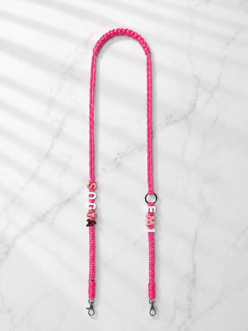 Op de afbeelding: Een felroze gevlochten koord met witte en roze letterkralen die SOCIA en EMI spellen. Het koord is voorzien van een zwarte ring en twee zilverkleurige metalen karabijnsluitingen aan de onderkant voor een veilige bevestiging van voorwerpen.