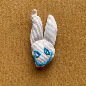 Peut inclure: Une décoration en forme de tête de lapin en tissu de toile texturée blanc cassé. Le lapin présente deux oreilles dressées et un visage brodé à la main avec du fil bleu vif formant deux yeux triangulaires et une large bouche souriante.