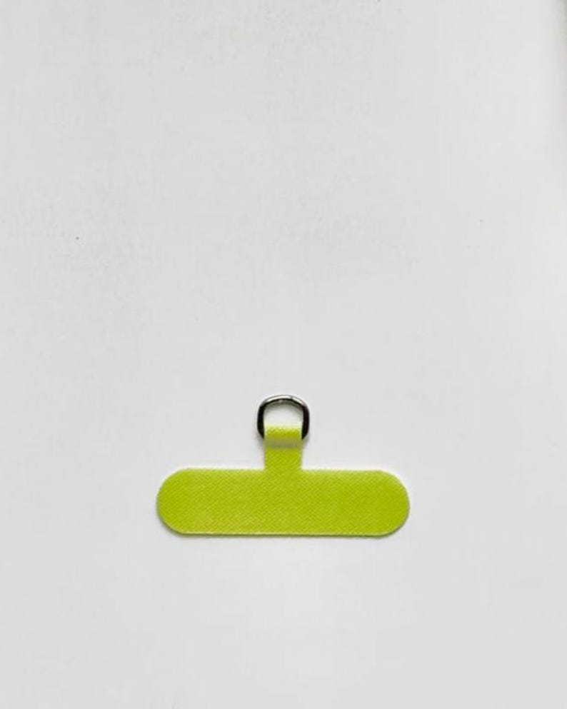Op de afbeelding: Een limoengroen, rechthoekig zelfklevend lipje met afgeronde uiteinden, voorzien van een kleine, zilverkleurige metalen D-ring in het midden aan de bovenkant. Het oppervlak van het lipje heeft een fijne, getextureerde afwerking voor bevestiging.