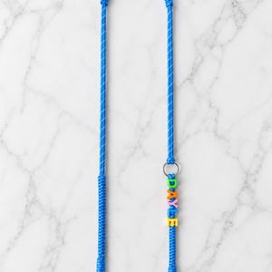 Op de afbeelding: Een felblauw gevlochten koord met een zilveren metalen ring en twee zilveren karabijnsluitingen aan de uiteinden. Het koord is versierd met kleurrijke plastic letterkralen die de naam DAYLE spellen in groen, oranje, roze, blauw en geel.