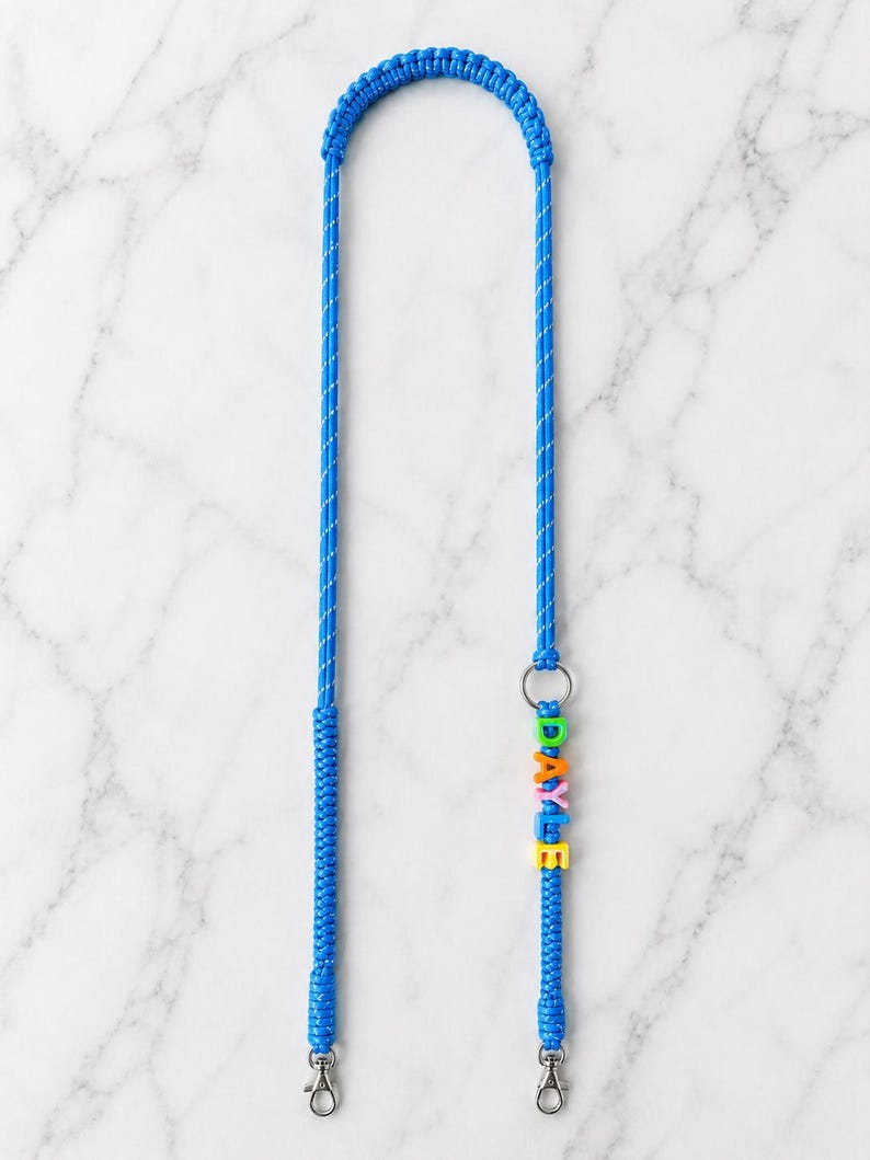Op de afbeelding: Een felblauw gevlochten koord met een zilveren metalen ring en twee zilveren karabijnsluitingen aan de uiteinden. Het koord is versierd met kleurrijke plastic letterkralen die de naam DAYLE spellen in groen, oranje, roze, blauw en geel.