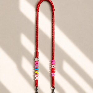 Op de afbeelding: Een rood gevlochten koord met twee zwarte metalen draaibare sluitingen aan de uiteinden. E&eacute;n kant is versierd met kleurrijke letterkralen die OLIVIA spellen, terwijl de andere kant roze letterkralen bevat die het woord MAMA vormen.