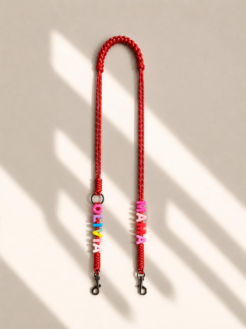 Op de afbeelding: Een rood gevlochten koord met twee zwarte metalen draaibare sluitingen aan de uiteinden. E&eacute;n kant is versierd met kleurrijke letterkralen die OLIVIA spellen, terwijl de andere kant roze letterkralen bevat die het woord MAMA vormen.