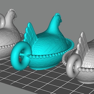 Könnte beinhalten: Drei keramikartige Behälter in Form von Hühnern, jeweils in einer anderen Farbe. Eine ist türkis, die anderen beiden sind grau. Sie sitzen in einem korbartigen Sockel mit Perlenrand. An jedem Huhn ist ein Ringgriff befestigt.