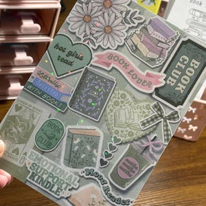 May include: A sticker sheet with book-related designs. The stickers feature daisies, books, hearts, and phrases such as "Book Lover" and "Emotional Support Kindle." The colour scheme is soft, with pastel pinks, greens, and purples.