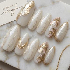 May include: A set of elegant, white artificial nails with gold leaf and floral accents. Some nails feature gold foil, while others have delicate gold lines or pink floral designs. A business card with the text "ORDER NAIL TIP vega" is also present.