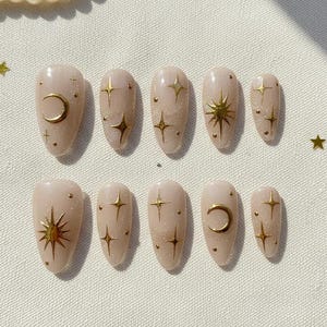 May include: A set of ten almond-shaped false nails in a nude color, adorned with gold celestial designs. The designs include crescent moons, stars, and starbursts. The nails are arranged on a white surface with scattered gold star-shaped confetti.