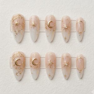 May include: A set of ten almond-shaped press-on nails featuring a soft white and nude gradient base. Each nail is decorated with gold metallic celestial motifs, including crescent moons, starbursts, and fine line work, accented with small clear rhinestones.
