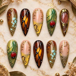 May include: A set of ten almond-shaped press-on nails featuring diverse designs including butterfly wing patterns, green foliage with 3D ladybug accents, gold filigree swirls on nude bases, and metallic gold frames with brown centers.