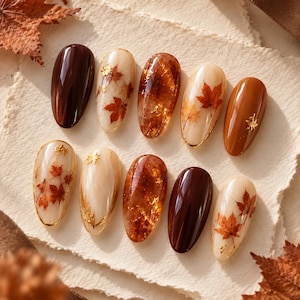 May include: A set of ten almond-shaped press-on nails featuring autumn-inspired designs. The collection includes solid dark brown and burnt orange nails, cream-colored nails with red maple leaf decals and gold foil accents, and amber-toned nails.