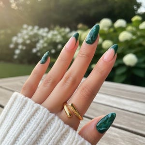 May include: A hand with long, almond-shaped nails painted with a green marble design and a dark green French tip. A gold ring adorns the finger. The nails are set against a blurred background of greenery and sunlight.