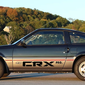 May include: A black and silver Honda CRX Pro car with a red and white stripe. The car is parked on a road with a backdrop of trees and a blue sky. The car has the text "CRX PRO" on the side.