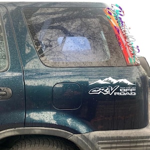 Peut inclure: Un SUV vert d'eau avec un autocollant blanc avec le texte "CRV OFF ROAD" et un graphique de chaîne de montagnes. Un autocollant coloré avec du texte est sur le cadre de la fenêtre. Le véhicule est à l'extérieur.