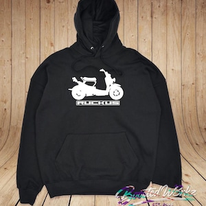 May include: Black hooded sweatshirt with a white graphic of a scooter and the word "RUCKUS" in a rectangular box. The sweatshirt has a front pocket and a drawstring hood. The background is a wooden wall.