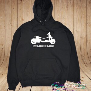 May include: Black hooded sweatshirt with a white graphic of a scooter and the word "RUCKUS" in a rectangular box. The sweatshirt has a front pocket and a drawstring hood. The bottom right corner has the text "Haunted Werks EST. 2025".