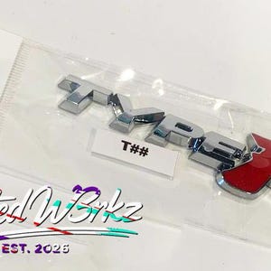 May include: Chrome and red "TYPE R" emblem in a clear plastic package. The emblem has a shiny chrome finish on the letters "TYPE" and a red "R". A small white label with "T##" is visible.