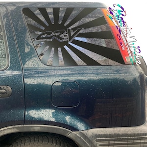 May include: A black and white vinyl decal applied to a car window. The design features the letters "CRV" in the center, with a rising sunburst pattern behind it. The car is dark teal.