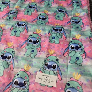 Puede incluir: Una manta estampada rosa, azul y verde con personajes de dibujos animados con trajes y sombreros verdes. La manta tiene una textura suave y afelpada y un patr&oacute;n repetitivo del personaje. La manta est&aacute; hecha a mano.