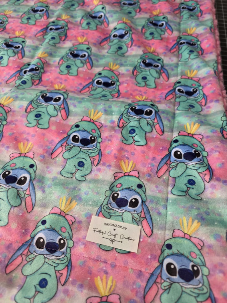 Puede incluir: Una manta estampada rosa, azul y verde con personajes de dibujos animados con trajes y sombreros verdes. La manta tiene una textura suave y afelpada y un patr&oacute;n repetitivo del personaje. La manta est&aacute; hecha a mano.