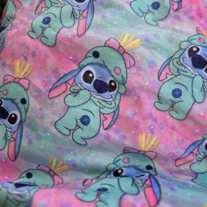 Puede incluir: Patr&oacute;n repetitivo del personaje Stitch de la pel&iacute;cula Lilo & Stitch. El personaje est&aacute; representado en un mono azul claro con un gorro a juego y detalles en rosa, sobre un fondo pastel rosa y azul.