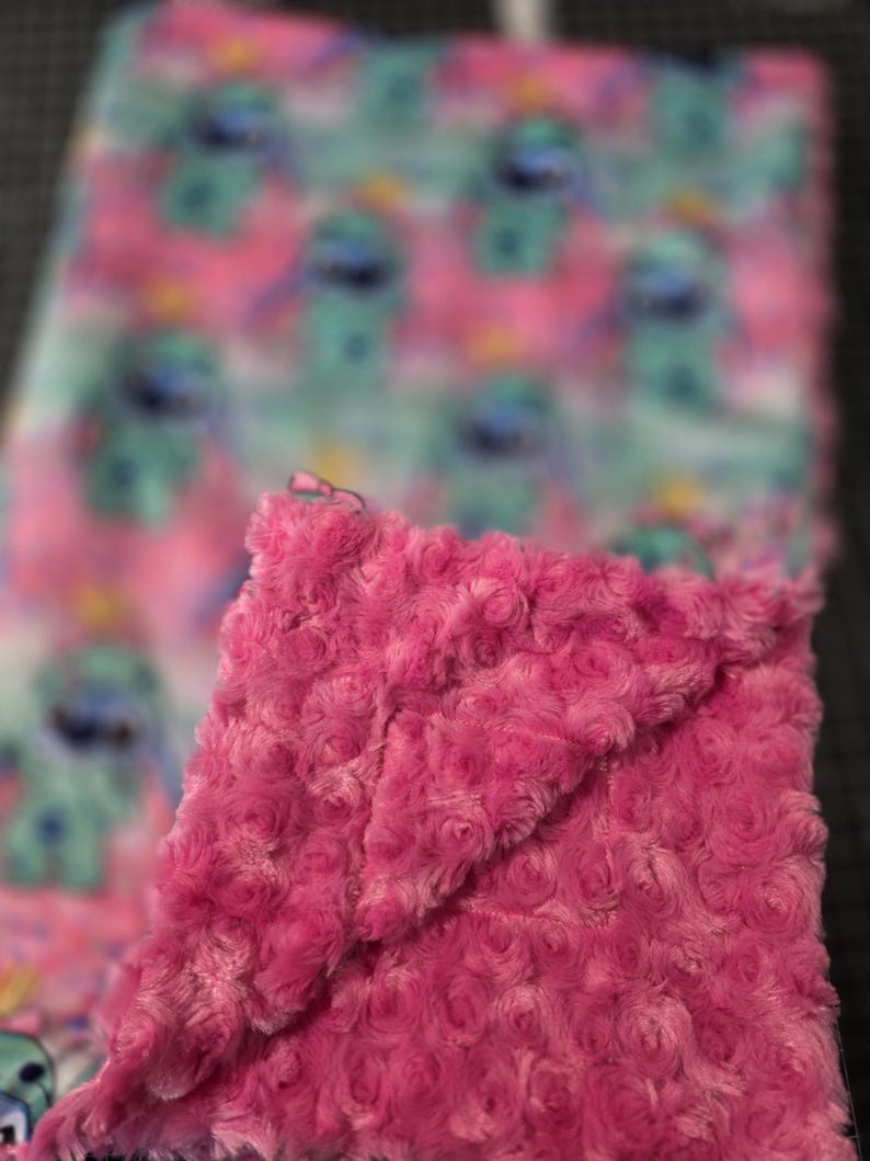 Puede incluir: Una manta rosa de felpa con una textura similar a una rosa est&aacute; doblada sobre una manta con un patr&oacute;n repetido de un personaje de dibujos animados azul sobre un fondo rosa y verde azulado. La manta es suave y acogedora.