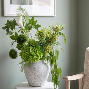May include: A vase of green flowers and foliage sits on a white pedestal. The vase is speckled grey. A framed abstract artwork hangs on the sage green wall. A cream-colored armchair with a blanket is in the background.