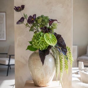 May include: A cream ceramic vase displaying a floral arrangement. The arrangement features dark purple flowers, green hydrangeas, and hanging green stems. The vase is placed on a light wood block, against a neutral-toned wall.