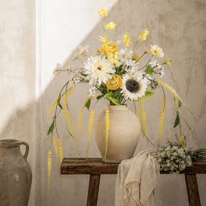 Könnte beinhalten: Ein Blumenarrangement mit weißen und gelben Blüten, darunter eine große Sonnenblume und Ranunkeln, in einer beigen Keramikvase. Die Vase steht auf einer dunklen Holzbank neben einem kleinen Strauß weißer Blumen und einem drapierten beigen Stoff.