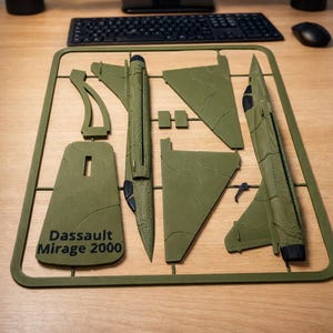 May include: An olive green Dassault Mirage 2000 model kit with detached parts, including wings and fuselage, sits on a wooden surface. A computer monitor displays an image of the aircraft in flight. The kit's frame includes the text "Dassault Mirage 2000".