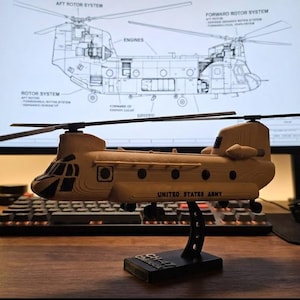 May include: A tan model of a United States Army Chinook helicopter, mounted on a black stand. The text "UNITED STATES ARMY" is visible on the side. A blueprint of the helicopter is in the background.
