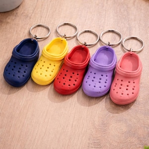 May include: A collection of five miniature clogs keychains in a row. The clogs are in various colors: navy blue, yellow, red, purple, and pink. Each keychain has a silver metal ring attached.