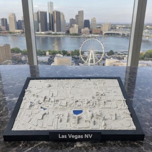 May include: A 3D model of Las Vegas, Nevada, in a black frame. The model is white with blue accents, depicting buildings and landmarks. The text "Las Vegas NV" is displayed on the frame. The background shows a cityscape and a Ferris wheel.
