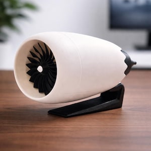 May include: A white and black model of a jet engine. The engine has a white cylindrical body with a black fan inside. It is mounted on a black stand. The model is on a wooden surface.