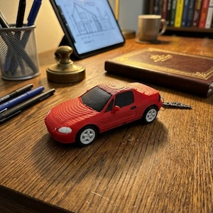 May include: A red toy car with white wheels sits on a wooden desk. The car has a detailed design, including windows and a roof. Other items on the desk include pens, a tablet displaying a blueprint, a book, and a coffee mug.