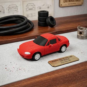 May include: A red 3D-printed Mazda Miata (NA/NB) model car, 1:64 scale, with white wheels. The model sits on a surface with a small gold plaque that reads "MAZDA MIATA (NA/NB) - 1:64 SCALE - 3D PRINTED ART".