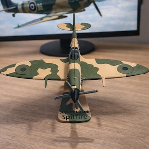 May include: A detailed model of a Supermarine Spitfire airplane, featuring a camouflage pattern of green and tan. The model has a propeller and is mounted on a stand with the name "Supermarine Spitfire" printed on it. The background includes a computer monitor.