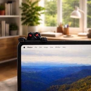 May include: A black laptop screen with a mountain landscape display. A black monster-like figure with red eyes is perched on the top edge of the laptop. The laptop is in an office setting with a window and desk in the background.
