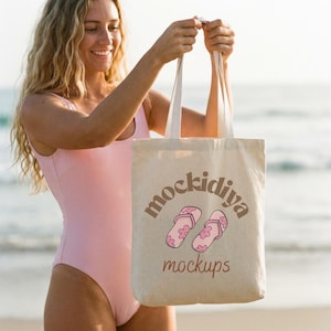 Op de afbeelding: Een beige draagtas met witte handvatten. De tas heeft de tekst "mockidiya mockups" en een afbeelding van roze slippers met bloemaccenten. De tas wordt vastgehouden door een persoon in een roze badpak.