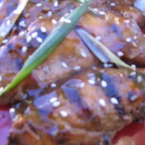 May include: Close-up of grilled chicken pieces glazed with a sweet and savory sauce, topped with sesame seeds and green onions. The chicken is arranged on a red plate.