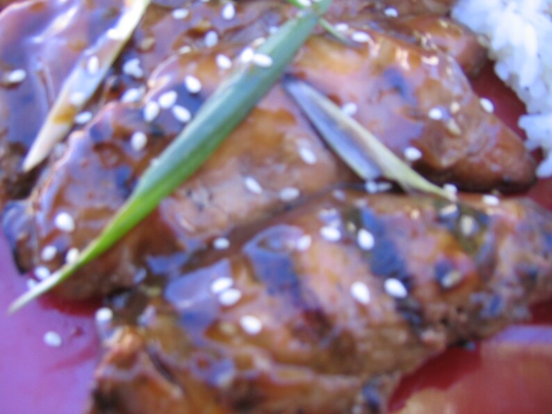 May include: Close-up of grilled chicken pieces glazed with a sweet and savory sauce, topped with sesame seeds and green onions. The chicken is arranged on a red plate.