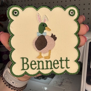May include: Embroidered patch with a scalloped edge, featuring a duck wearing bunny ears and a bow tie. The duck is brown, pink, and green. The name "Bennett" is embroidered in green below the duck. The patch is approximately 4 inches square.